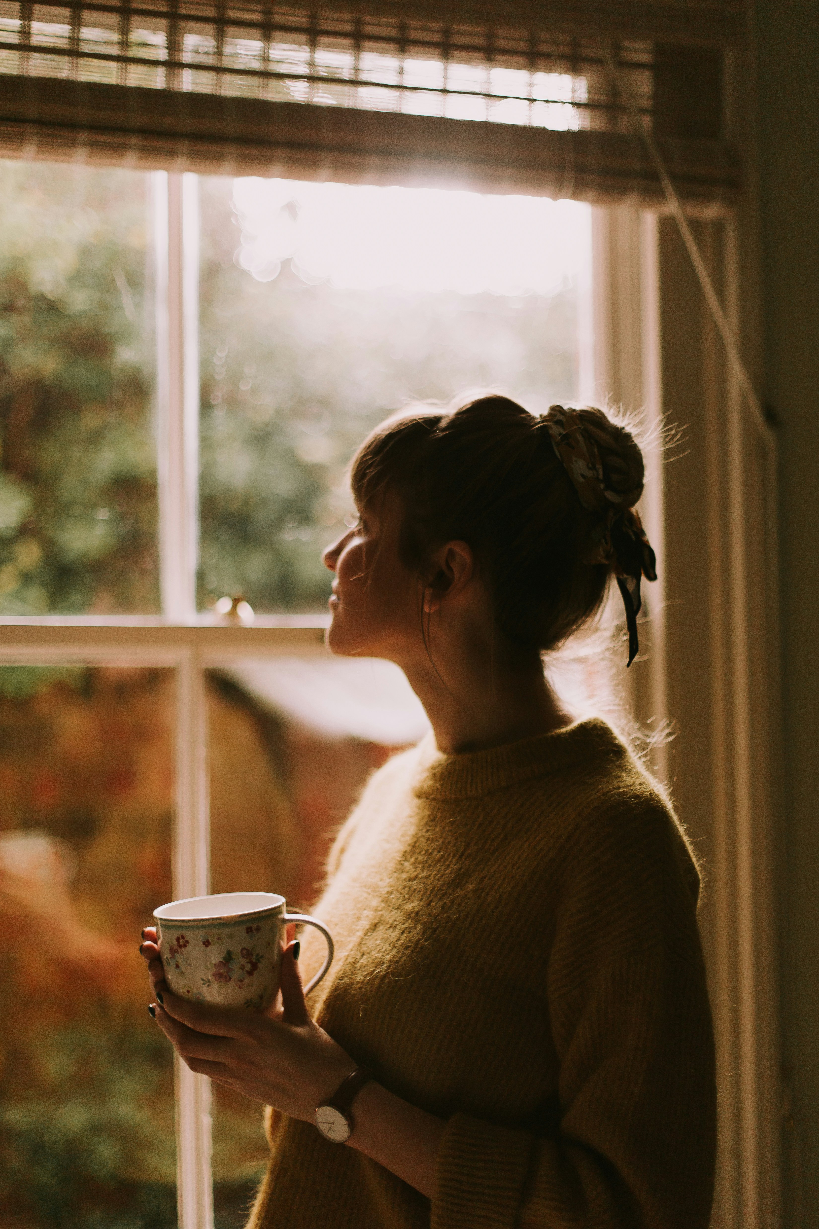 A woman pauses by a window holding a warm mug in afternoon light